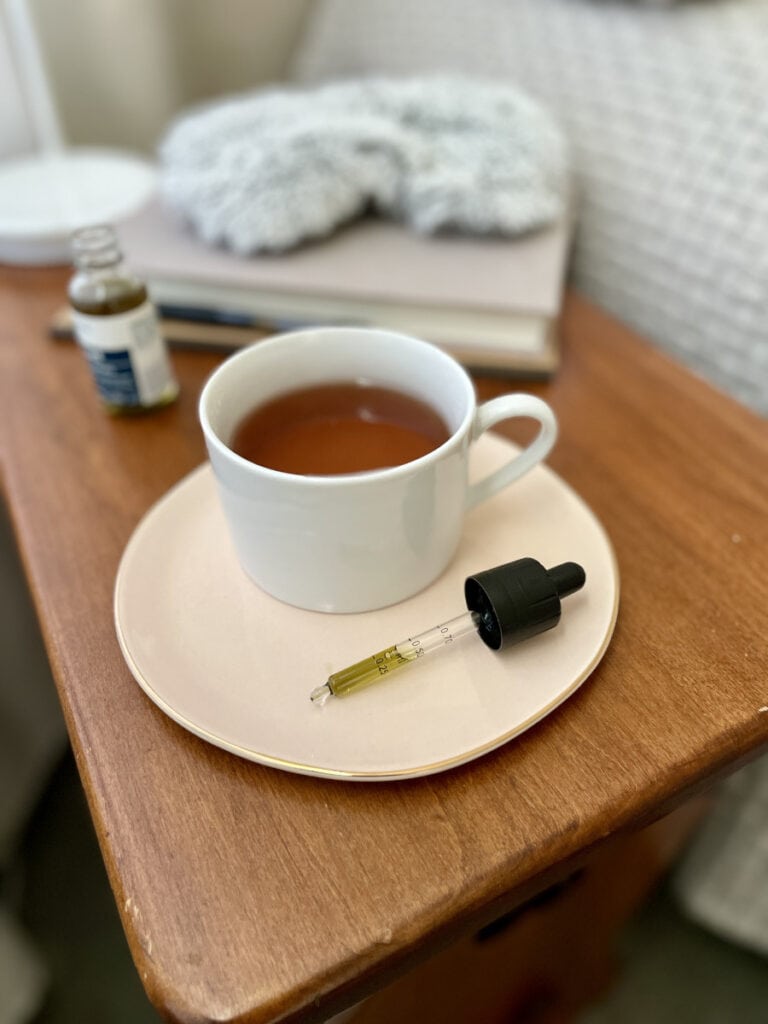 A dropper of CBD oil sits next to a cup of tea on a wooden beside table - Annie's CBD Guide